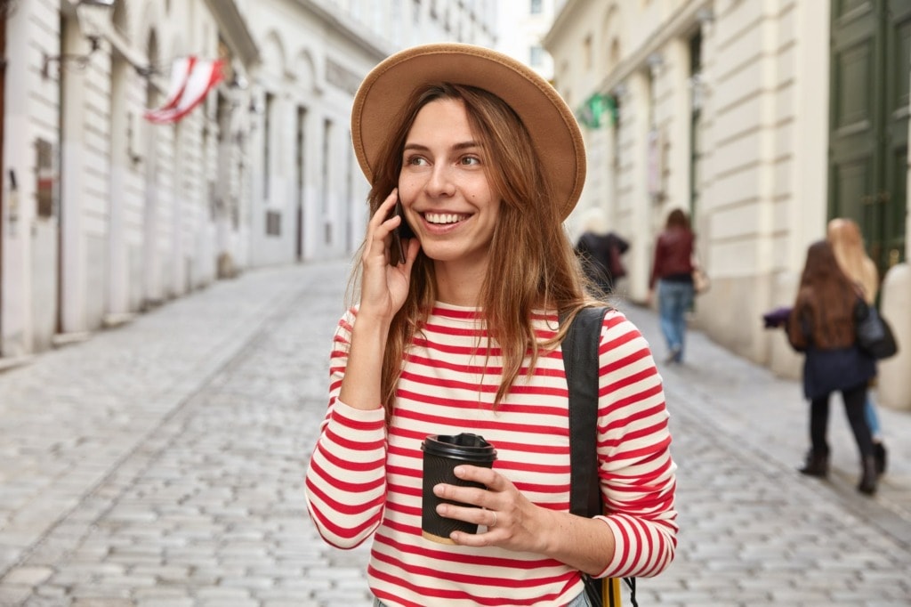 gorgeous smiling tourist strolls around city has phone conversation holds takeaway coffee focused somewhere into distance min