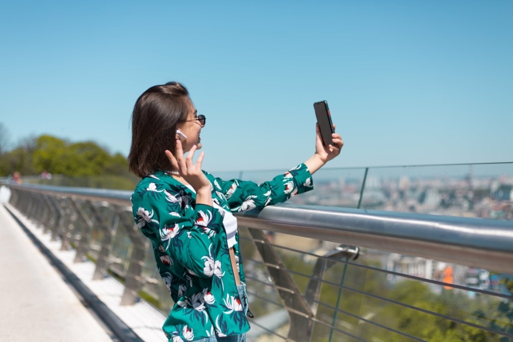outdoor portrait woman casual green shirt sunny day stands bridge looking phone screen take selfie make video call wireless bluetooth headphones ears min