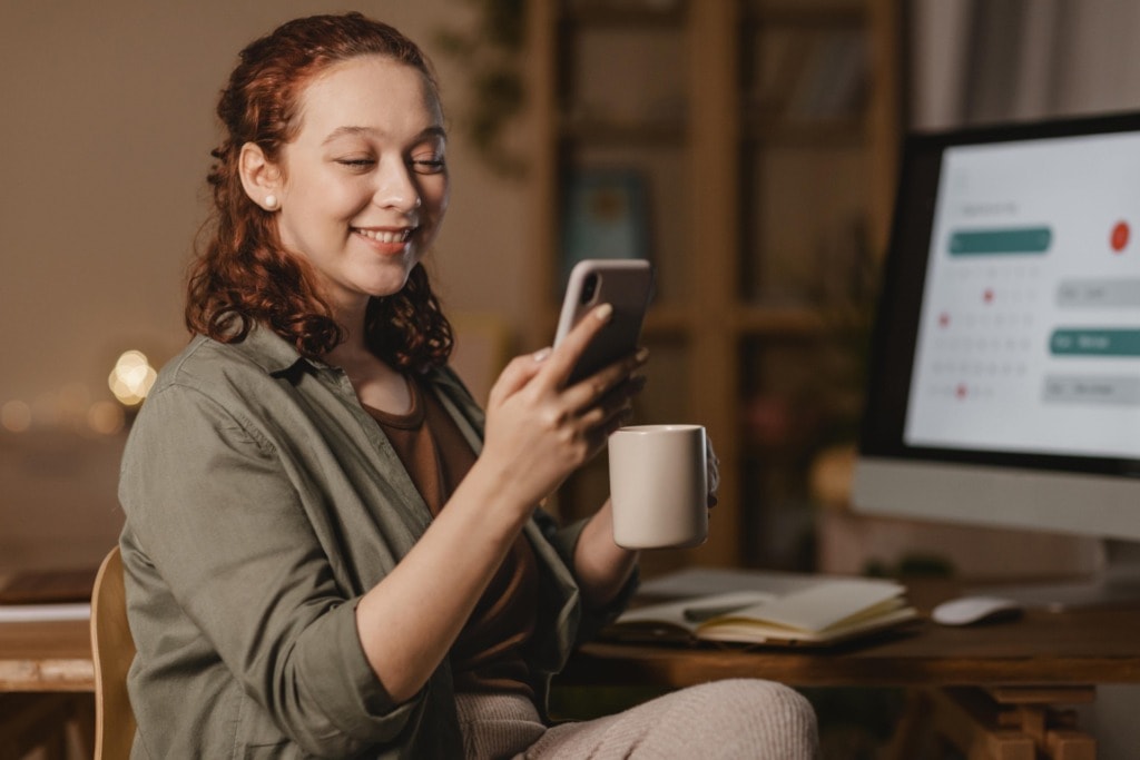 woman home using smartphone front computer while having coffee min